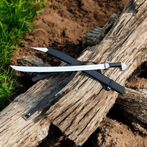 May include: A silver-bladed sword with a black handle and sheath, lying on a weathered wooden log. The sword features a curved blade and a decorative hilt. A black leather strap is also present.