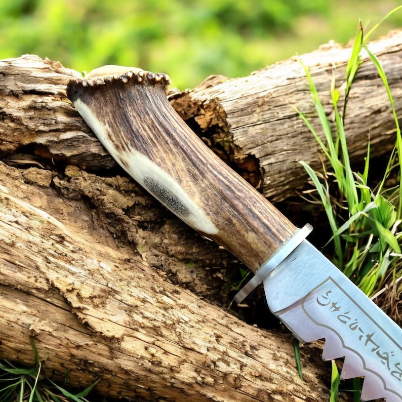 May include: Close-up of a hunting knife with a natural antler handle and a serrated blade. The blade has etched text. The knife rests on a weathered log, with green grass in the background.