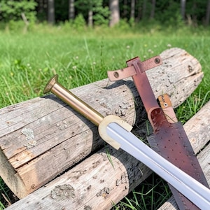 May include: A double-edged sword with a polished brass pommel and hilt, featuring a straight steel blade. Beside the sword lies a dark brown leather scabbard with a textured surface, metal rivets, and a leather strap with two metal snap buttons.