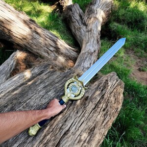 May include: A toy sword with a light blue blade and a gold-colored hilt. The hilt has a decorative design with a blue center. The sword is held against a weathered wooden log with green grass in the background.