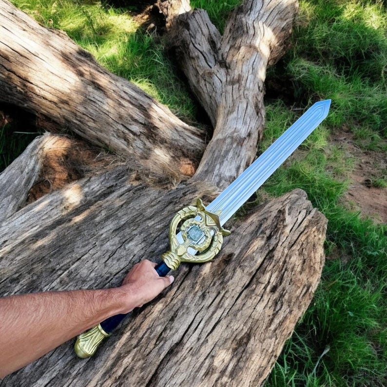 May include: A toy sword with a light blue blade and a gold-colored hilt. The hilt has a decorative design with a blue center. The sword is held against a weathered wooden log with green grass in the background.