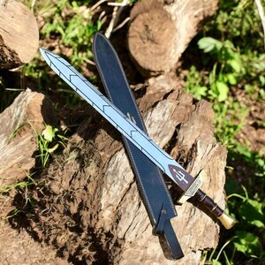 May include: A decorative sword with a light blue blade featuring a geometric design, resting on a black sheath. The hilt is brown with a gold-colored pommel. The sword is displayed on a weathered wooden surface.