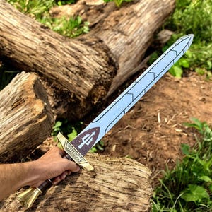 May include: A toy sword with a light blue blade and a gold hilt. The hilt features a trident symbol and the words "WARRIOR'S EDGE". The sword is held against a backdrop of wood and greenery.
