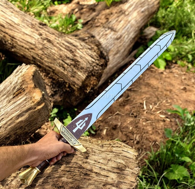 May include: A toy sword with a light blue blade and a gold hilt. The hilt features a trident symbol and the words "WARRIOR'S EDGE". The sword is held against a backdrop of wood and greenery.