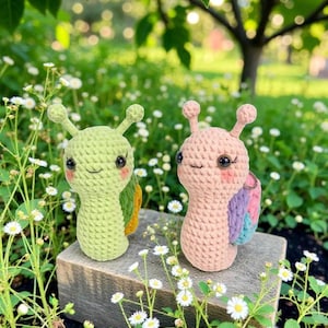 Könnte beinhalten: Zwei gehäkelte Schneckenfiguren. Eine ist hellgrün, die andere rosa. Beide haben lächelnde Gesichter, Antennen und farbenfrohe Muscheldetails. Sie werden auf einem Holzblock ausgestellt, umgeben von Grün und kleinen weißen Blumen.