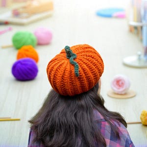 Könnte beinhalten: Ein orangefarbener gehäkelter Kürbishut mit grünem Stiel wird von einer Person getragen. Der Hut ist aus dickem Garn gefertigt und hat gerippte Details. Bunte Wollknäuel in Grün, Rosa, Orange und Lila sind im Hintergrund. Die Person trägt ein kariertes Hemd.
