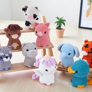 Könnte beinhalten: Eine Sammlung handgefertigter gehäkelter Tierfiguren, darunter eine Kuh, ein Schwein, ein Waschbär, ein Nilpferd, ein Einhorn, ein Elefant, ein Tiger und eine Ente. Die Figuren sind auf einem Holzregal ausgestellt.