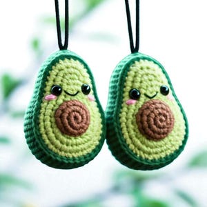 Könnte beinhalten: Zwei handgefertigte Avocado-Ornamente aus Häkelarbeit mit lächelnden Gesichtern, schwarzen Augen und rosigen Wangen. Die Avocados sind grün mit einem braunen Kern und werden von schwarzen Kordeln gehalten. Diese niedlichen Ornamente sind perfekt für eine Küche oder als Geschenk.