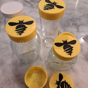 May include: Several glass jars with yellow lids featuring a black bee design. One lid is off, revealing the jar's opening. The jars vary in shape and size, with a white lid jar in the background. The jars are on a marble surface.