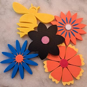 May include: A collection of colorful, 3D-printed floral and insect decorations. The image features a yellow bee, a black flower with a pink center, and several flowers in shades of blue, orange, and yellow. The decorations are arranged on a light-colored surface.