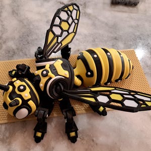 May include: A detailed, 3D-printed model of a bee, featuring a yellow and black color scheme. The bee has segmented body parts, including a rounded abdomen, and intricate wing designs with white and black accents. The model is placed on a textured, tan surface.