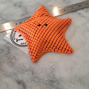 May include: An orange, five-pointed star-shaped object with a textured, grid-like surface and two small black eyes. A metal ruler is positioned behind the object, showing it measures approximately 4.5 inches across at its widest point.