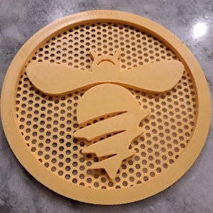 May include: A round, yellow coaster featuring a bee design. The bee is in the center, with a honeycomb pattern in the background. The coaster has a raised edge.