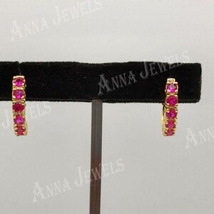 May include: A pair of gold-colored hoop earrings, each adorned with a row of vibrant red gemstones. The earrings are displayed on a black velvet stand against a neutral background. The brand name "Anna Jewels" is visible.