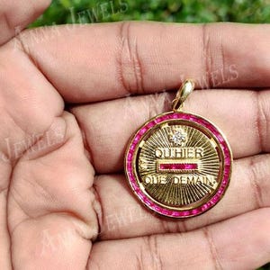 May include: A gold-colored pendant with a circular design, featuring a ring of red gemstones around the edge. The center has text that reads "OUTHIER ONE DEMAIN". The pendant is held in a hand.