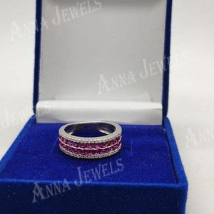 May include: A silver ring with a band of small, vibrant red gemstones, framed by rows of smaller, clear stones. The ring is displayed in a blue velvet jewelry box with the text "ANNA JEWELS".