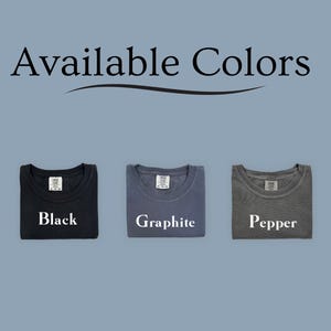 Puede incluir: La imagen muestra tres camisetas dobladas en colores negro, grafito y pimienta. El texto "Available Colors" est&aacute; en la parte superior. Cada camiseta tiene una etiqueta y el nombre del color impreso debajo.