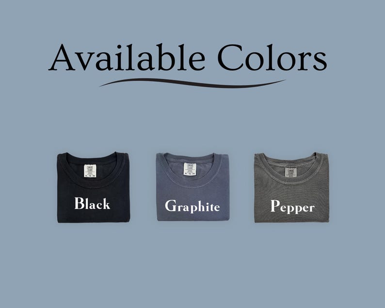 Puede incluir: La imagen muestra tres camisetas dobladas en colores negro, grafito y pimienta. El texto "Available Colors" est&aacute; en la parte superior. Cada camiseta tiene una etiqueta y el nombre del color impreso debajo.