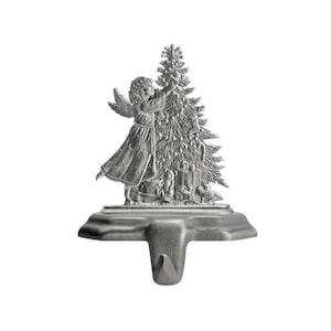 May include: Silver-colored Christmas stocking holder featuring an angel decorating a Christmas tree. The angel has wings and is wearing a long dress. The tree is decorated with ornaments and presents are at the base. The holder has a hook for hanging.