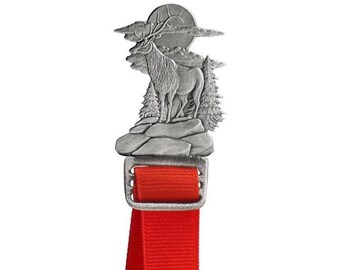 Puede incluir: Colgante de metal plateado que representa un alce sobre una superficie rocosa, con una luna llena y nubes en el fondo. El colgante está sujeto a una cinta roja. El diseño tiene temática de la naturaleza.