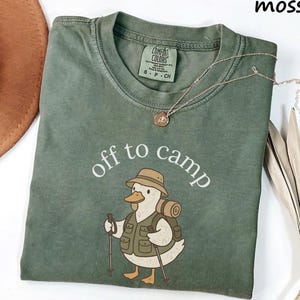 May include: A moss green t-shirt with the words "off to camp" and a cartoon duck wearing a hat, backpack, and vest. A gold necklace with a pendant is on the shirt. A brown hat is in the upper left corner.