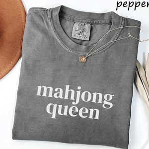 May include: A gray, short-sleeved t-shirt with the text "mahjong queen" in white. The shirt has a crew neck and a small tag near the collar. A gold necklace with a pendant is draped across the shirt. The word "pepper" is in the top right corner.
