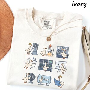 May include: Ivory-colored t-shirt featuring cartoon geese in various engineering and space-related scenarios. The geese are depicted with tools, rockets, and blueprints. The shirt has a Comfort Colors label.