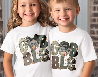 May include: Two children wearing white t-shirts with a Thanksgiving-themed design. The shirts feature the word "GOBBLE" in a mix of camouflage and patterned letters, with a turkey graphic and fall leaves. One shirt has a bow, the other a cap.