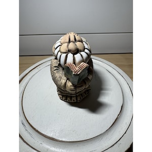 May include: A ceramic figurine of a bearded man reading a book. The figure has a tan face and beard with white accents, wearing a brown robe. The book is open, with green and brown pages. The figurine is on a white, distressed, round surface.