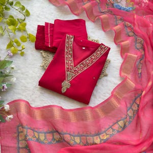 Puede incluir: Un vibrante conjunto de salwar kameez indio rojo. La parte superior presenta intrincados bordados dorados a lo largo del escote y un diseño bordado dorado a juego en el centro. El conjunto incluye pantalones rojos a juego y una dupatta rosa transparente con bordes dorados y azules.