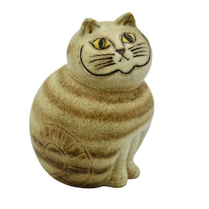 May include: A ceramic cat figurine in a seated position. The cat is tan with brown stripes and black facial details. The eyes are yellow. The figurine has a detailed design on the lower back. The website address "www.coscombefineporcelain.com" is at the top.