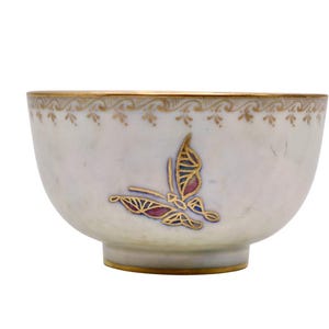 May include: A white ceramic bowl with a gold rim and a butterfly design. The butterfly has gold outlines with red and blue accents. The bowl has a decorative gold pattern around the rim and a gold base.