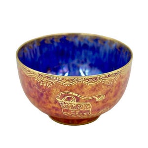 May include: A decorative porcelain bowl with a deep blue interior and a reddish-brown exterior. The rim and exterior feature gold detailing, including a basket design. The website address "www.coscombefineporcelain.com" is visible at the top.