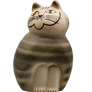 Könnte beinhalten: Keramik-Katzenfigur mit lächelndem Gesicht, gelben Augen und schwarzen Schnurrhaaren. Die Katze hat ein gestreiftes Muster in Braun- und Beigetönen. Die Figur sitzt aufrecht, die Pfoten sind sichtbar.