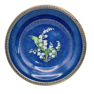 May include: A round, blue porcelain plate with a gold rim and a floral design. The plate features a central image of white lily of the valley flowers with green leaves. The rim has a gold pattern. The website address "www.coscombefineporcelain.com" is at the top.