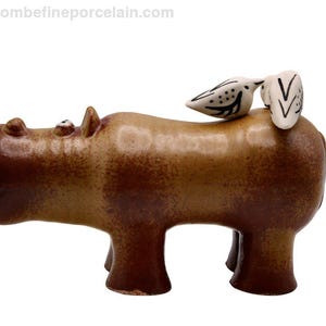 Könnte beinhalten: Eine braune Keramik-Nilpferdfigur mit zwei kleinen weißen Vögeln mit schwarzen Markierungen auf dem Rücken. Die Figur hat eine glänzende Oberfläche. Der Hintergrund ist weiß.