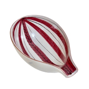 May include: A white porcelain dish with a red and black striped pattern. The dish has a unique shape, with a rounded top and a narrow, elongated base. The design includes a central leaf-like motif.