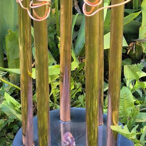 May include: A copper and brass sculpture featuring six vertical tubes, linked by copper wire, on a dark blue base. A purple crystal is in the centre. The sculpture is outdoors, surrounded by green foliage.