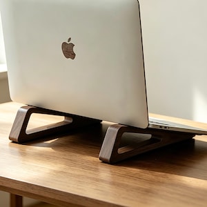 Può includere: Un computer portatile color argento con il logo Apple sul coperchio è posizionato su un supporto in legno marrone scuro. Il supporto presenta un design geometrico a telaio aperto con supporti angolati che sollevano il portatile.