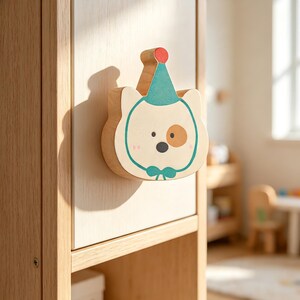 Puede incluir: Un pomo de mueble de madera clara con forma de cabeza de oso que lleva un sombrero de fiesta azul turquesa con un pompón rojo. La cara del oso tiene una mancha marrón sobre un ojo, una nariz negra y una pajarita turquesa en la madera.