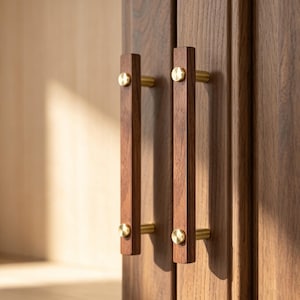 May include: Two vertical cabinet handles made of dark brown wood with a visible grain pattern. Each handle is attached to the door with two cylindrical brass-colored metal posts that feature flat, circular screw heads on the front surface.
