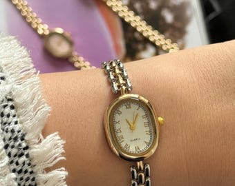 May include: Gold-toned quartz watch with an oval face and Roman numeral hour markers. The watch has a gold bezel and a two-tone gold and silver band. The watch is on a wrist, with two other watches visible in the background.