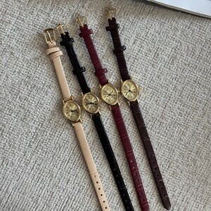 K&ouml;nnte beinhalten: Vier elegante Armbanduhren mit goldfarbenen ovalen Zifferbl&auml;ttern und r&ouml;mischen Ziffern. Die Armb&auml;nder sind in Creme, Schwarz, Burgunderrot und Braun gehalten, mit passenden goldenen Schnallen. Die Uhren liegen auf einer strukturierten, hellgrauen Oberfl&auml;che.