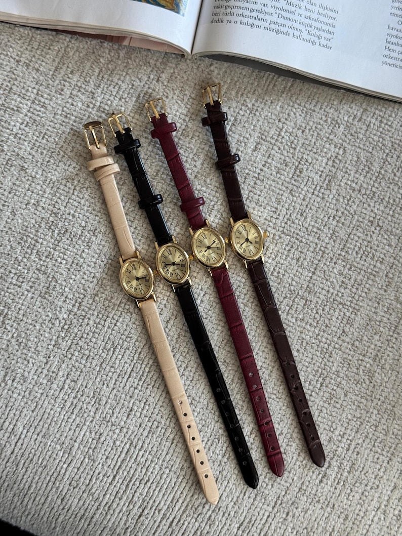K&ouml;nnte beinhalten: Vier elegante Armbanduhren mit goldfarbenen ovalen Zifferbl&auml;ttern und r&ouml;mischen Ziffern. Die Armb&auml;nder sind in Creme, Schwarz, Burgunderrot und Braun gehalten, mit passenden goldenen Schnallen. Die Uhren liegen auf einer strukturierten, hellgrauen Oberfl&auml;che.