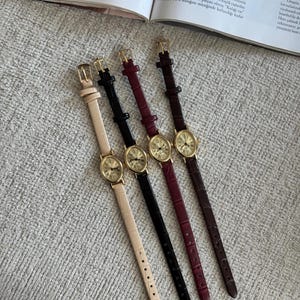 K&ouml;nnte beinhalten: Vier Damenuhren mit goldfarbenen Geh&auml;usen und Lederarmb&auml;ndern in Beige, Schwarz, Burgunderrot und Braun. Die Uhren haben ein ovales Zifferblatt mit r&ouml;mischen Ziffern und eine Schnalle.