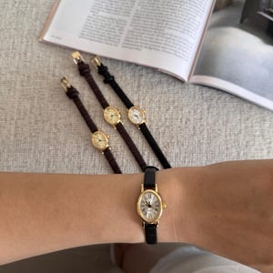K&ouml;nnte beinhalten: Eine Sammlung goldfarbener Armbanduhren mit ovalen Zifferbl&auml;ttern und r&ouml;mischen Ziffern. Die Uhren haben Lederarmb&auml;nder in Schwarz und Braun. Eine Uhr wird am Handgelenk getragen. Im Hintergrund ist eine Zeitschrift ge&ouml;ffnet.