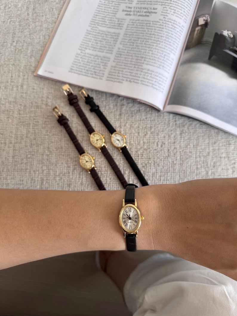 K&ouml;nnte beinhalten: Eine Sammlung goldfarbener Armbanduhren mit ovalen Zifferbl&auml;ttern und r&ouml;mischen Ziffern. Die Uhren haben Lederarmb&auml;nder in Schwarz und Braun. Eine Uhr wird am Handgelenk getragen. Im Hintergrund ist eine Zeitschrift ge&ouml;ffnet.