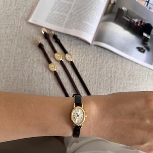 K&ouml;nnte beinhalten: Eine goldfarbene Armbanduhr mit schwarzem Lederarmband wird am Handgelenk getragen. Die Uhr hat ein wei&szlig;es Zifferblatt mit r&ouml;mischen Ziffern. Drei weitere &auml;hnliche Uhren mit unterschiedlich farbigen Armb&auml;ndern sind ebenfalls sichtbar.