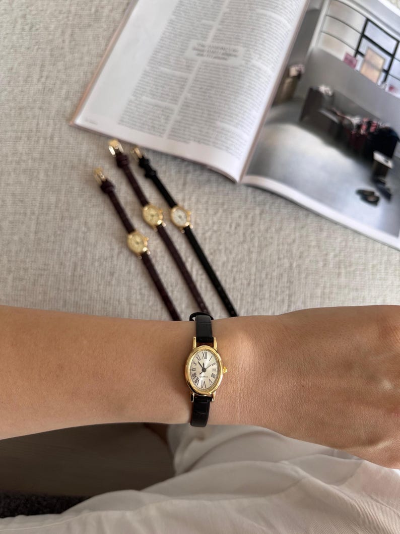 K&ouml;nnte beinhalten: Eine goldfarbene Armbanduhr mit schwarzem Lederarmband wird am Handgelenk getragen. Die Uhr hat ein wei&szlig;es Zifferblatt mit r&ouml;mischen Ziffern. Drei weitere &auml;hnliche Uhren mit unterschiedlich farbigen Armb&auml;ndern sind ebenfalls sichtbar.