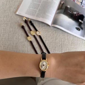 K&ouml;nnte beinhalten: Eine goldfarbene Armbanduhr mit schwarzem Lederarmband wird am Handgelenk getragen. Die Uhr hat ein wei&szlig;es Zifferblatt mit r&ouml;mischen Ziffern. Drei weitere Uhren mit goldenen Zifferbl&auml;ttern und Lederarmb&auml;ndern sind im Hintergrund zu sehen.